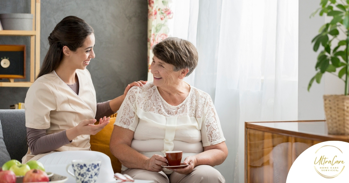 Smiling caregiver with her hand on an older woman’s shoulder as they share tea on the couch, showing home care vs assisted living